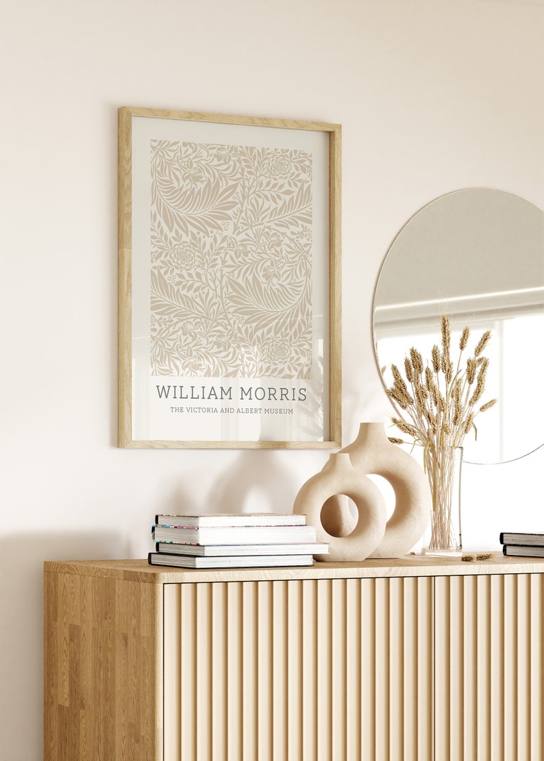 May include: A framed print of a beige floral pattern with the text "WILLIAM MORRIS THE VICTORIA AND ALBERT MUSEUM" at the bottom. The print is hanging on a white wall above a wooden cabinet with a round mirror and a vase with dried flowers.