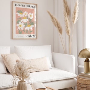 May include: A white couch with two pillows and a vase of dried flowers on a coffee table. A framed print with the text "FLOWER MARKET" and "THE FLORAL GALLERY LONDON" hangs on the wall. A tall vase with dried pampas grass stands in the corner.