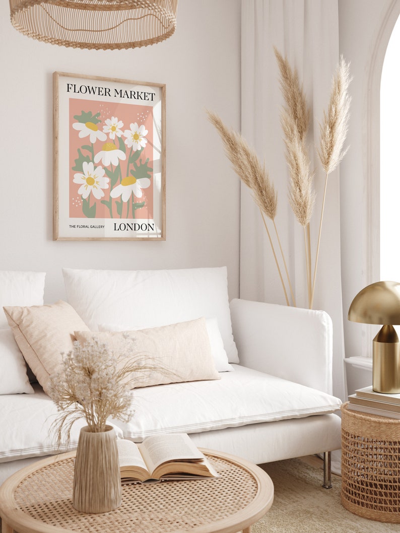 May include: A white couch with two pillows and a vase of dried flowers on a coffee table. A framed print with the text "FLOWER MARKET" and "THE FLORAL GALLERY LONDON" hangs on the wall. A tall vase with dried pampas grass stands in the corner.