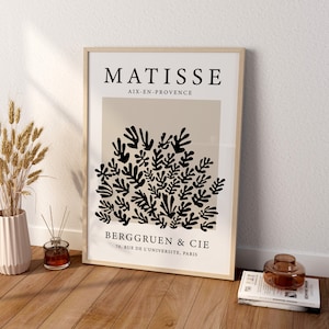 May include: A framed print of a black and white abstract art piece by Henri Matisse. The print features a cluster of stylized leaves and branches on a beige background. The text "MATISSE" is at the top of the print, followed by "AIX-EN-PROVENCE". Below the artwork, the text "BERGGRUEN & CIE" and "70, RUE DE L'UNIVERSITE, PARIS" are printed.