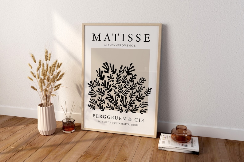 May include: A framed print of a black and white abstract art piece by Henri Matisse. The print features a cluster of stylized leaves and branches on a beige background. The text "MATISSE" is at the top of the print, followed by "AIX-EN-PROVENCE". Below the artwork, the text "BERGGRUEN & CIE" and "70, RUE DE L'UNIVERSITE, PARIS" are printed.