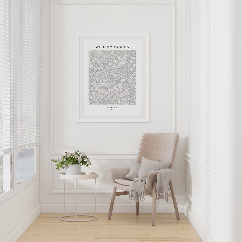 May include: A framed William Morris "Larkspur 1875" print hangs on a white wall. A beige armchair with a throw and pillow sits nearby. A small, round side table holds a potted plant. The room has a window with blinds and light wood flooring.