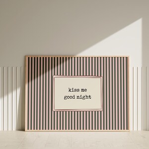 Puede incluir: Un estampado enmarcado con un fondo a rayas rosas y negras y un centro color crema. El centro presenta el texto "kiss me good night" en una fuente sencilla. El marco es de color madera clara.