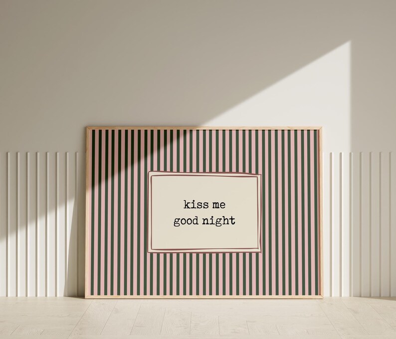 Puede incluir: Un estampado enmarcado con un fondo a rayas rosas y negras y un centro color crema. El centro presenta el texto "kiss me good night" en una fuente sencilla. El marco es de color madera clara.