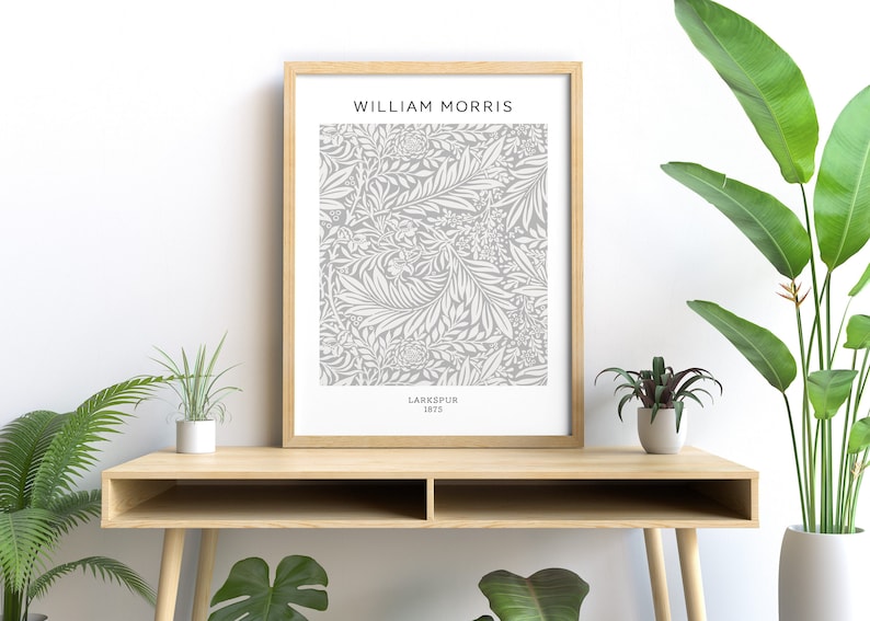 May include: A framed print featuring a gray and white floral pattern, labeled "Larkspur 1875" by William Morris. The artwork is displayed on a light wood desk, with green plants in the foreground.