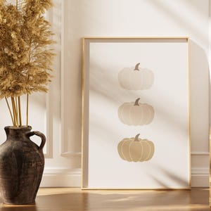 May include: A framed print with three pumpkin illustrations in beige and brown tones. The pumpkins are stacked on a cream backdrop, enclosed in a gold frame. The artwork is positioned near a decorative vase, adding to the autumnal theme.