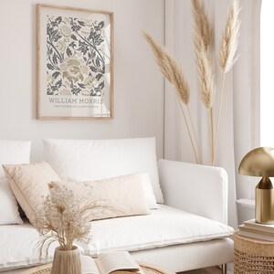 May include: A white couch with two pillows and a vase of dried flowers on a coffee table. A framed print of a floral design by William Morris hangs on the wall behind the couch. A tall vase of dried pampas grass stands in the corner of the room.