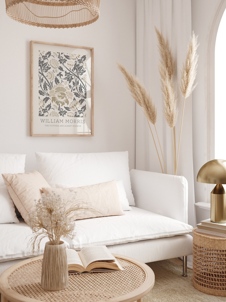 May include: A white couch with two pillows and a vase of dried flowers on a coffee table. A framed print of a floral design by William Morris hangs on the wall behind the couch. A tall vase of dried pampas grass stands in the corner of the room.