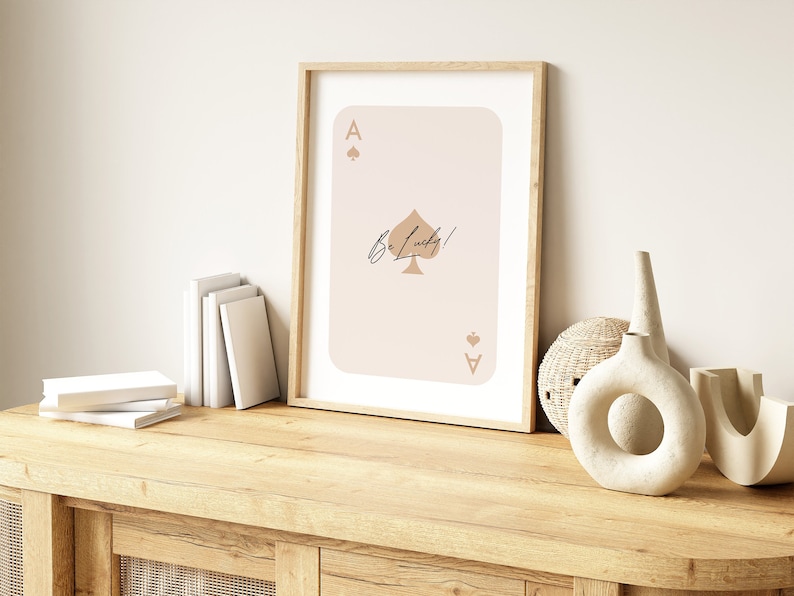 May include: A framed print of a playing card with a spade design and the text "Be Lucky!" in a cursive font. The card is a light pink color with a brown spade and the number "A" in the top left and bottom right corners.