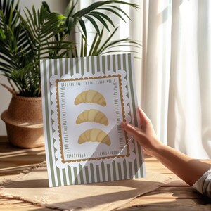 May include: A print depicting three croissants within a decorative frame. The frame is set against a backdrop of green and white vertical stripes. The print is held in front of a window, with a plant visible in the background.
