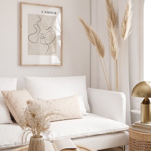 May include: A white sofa with a beige throw pillow and a vase of dried flowers on a coffee table. A framed print with the text "L'AMOUR" hangs on the wall above the sofa. Dried pampas grass is in a vase on the floor next to the sofa.