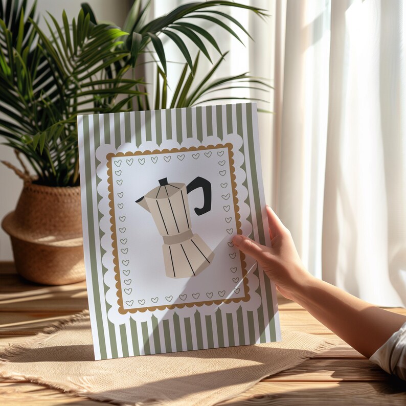 Peut inclure: Une impression d&eacute;corative repr&eacute;sentant une illustration de cafeti&egrave;re. L'impression a un fond blanc avec une bordure marron et un bord festonn&eacute;. Le fond pr&eacute;sente des rayures verticales vertes et blanches. L'impression est tenue par une personne.