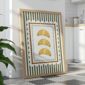 May include: A framed print featuring three golden croissants on a white background with a decorative border. The background has vertical green and white stripes. The frame is made of light-coloured wood.
