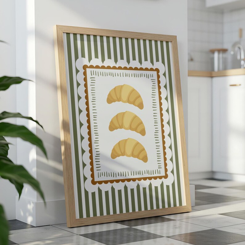 May include: A framed print featuring three golden croissants on a white background with a decorative border. The background has vertical green and white stripes. The frame is made of light-coloured wood.