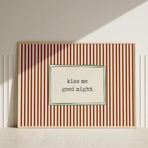 Puede incluir: Un cuadro enmarcado con un marco de madera clara. La obra presenta un rect&aacute;ngulo color crema con las palabras "kiss me good night" en texto negro, sobre un fondo de rayas verticales marrones y crema.