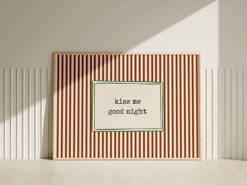 Puede incluir: Un cuadro enmarcado con un marco de madera clara. La obra presenta un rect&aacute;ngulo color crema con las palabras "kiss me good night" en texto negro, sobre un fondo de rayas verticales marrones y crema.