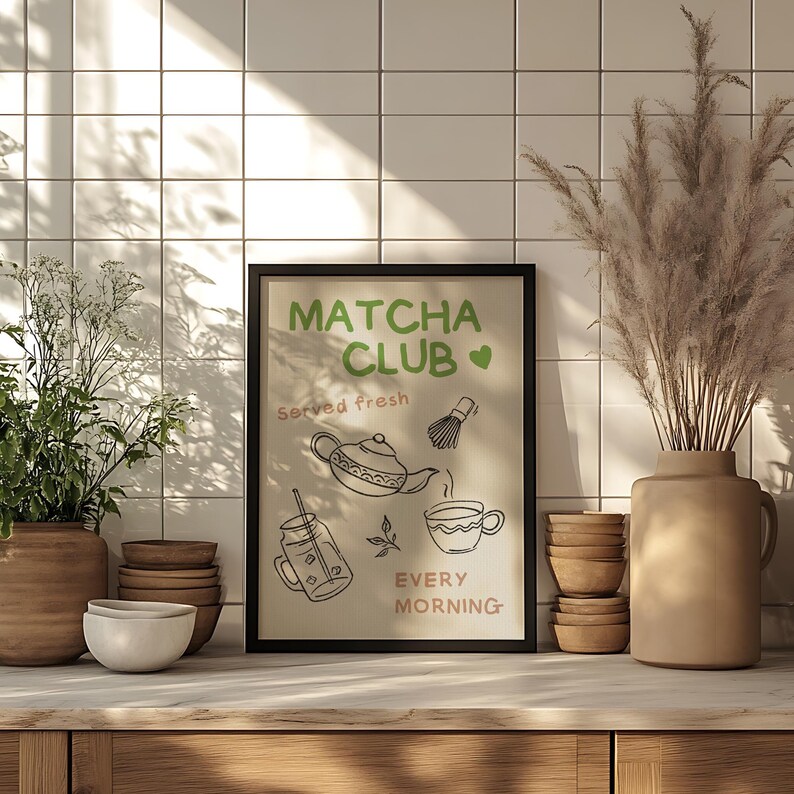 Pu&ograve; includere: Un poster incorniciato con la scritta 'Matcha Club' in verde e illustrazioni a tema t&egrave;. Il poster &egrave; su una superficie di legno, con un vaso di piante secche, ciotole e una pianta in vaso sullo sfondo.