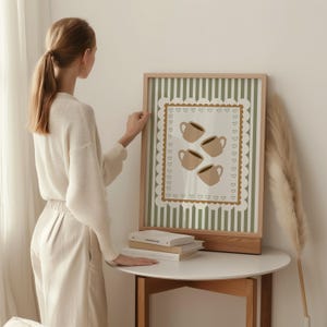May include: A framed art print featuring four coffee cups in a beige color, set against a striped green and white background. The print is framed in a light wood frame and rests on a white table.