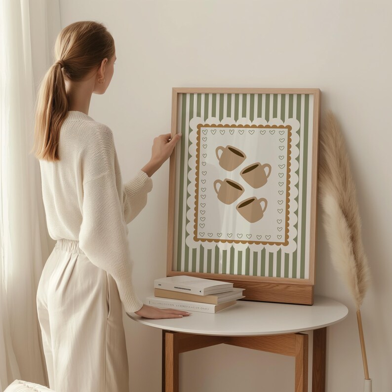 May include: A framed art print featuring four coffee cups in a beige color, set against a striped green and white background. The print is framed in a light wood frame and rests on a white table.