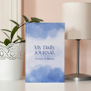 Puede incluir: Un diario de tapa dura con un diseño de nubes azules en acuarela. La portada dice "My Daily Journal" con el nombre "Emma Williams" debajo. El diario se muestra sobre una superficie blanca junto a una planta en maceta y una lámpara.