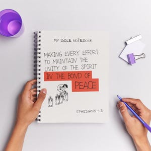 Puede incluir: Un cuaderno en espiral blanco con el texto "MY BIBLE NOTEBOOK" y una cita sobre la unidad y la paz. Una mano sostiene un bolígrafo azul, y también se ven una taza morada y un clip. El cuaderno está abierto.