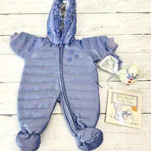 May include: A pale blue baby snowsuit with a faux fur-trimmed hood. The snowsuit features a zip and snowflake embroidery. A small teddy bear and a book titled "Bitty Bear's Frosty Fun" are also visible.