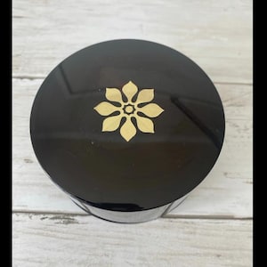Puede incluir: Un recipiente redondo negro con un acabado brillante. La tapa presenta un diseño floral dorado en el centro. El recipiente está sobre una superficie de madera clara.