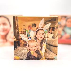 May include: A wooden block featuring a photo of two children wearing bunny ears. The older child is wearing a yellow shirt, and the younger child is wearing a black shirt and holding a yellow egg. The background is a blurred interior.