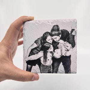Peut inclure: Photographie en noir et blanc d'une famille de quatre personnes. Un homme, une femme et deux enfants sourient et se regardent. L'homme tient le plus jeune enfant en l'air. La photo est imprimée sur un dessous de verre carré en pierre blanche.