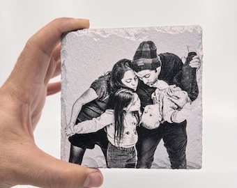 May include: Black and white photo of a family of four. A man, woman, and two children are smiling and looking at each other. The man is holding the youngest child in the air. The photo is printed on a square, white stone coaster.