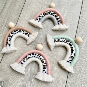 May include: Four handmade macrame rainbow decorations. Each rainbow features a wooden bead, a leopard print fabric layer, and layers of pink, grey, white, and green yarn. The rainbows have a fringed bottom.