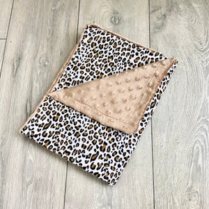 May include: A folded baby blanket with a leopard print pattern in black, brown, and tan on a white background. The blanket has a tan, textured border. The blanket is folded on a light grey wooden surface.
