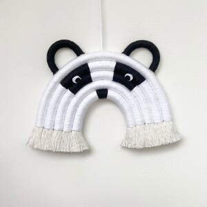 Puede incluir: Colgante de pared de arco&iacute;ris de macram&eacute; blanco y negro con un dise&ntilde;o de cara de tej&oacute;n. El arco&iacute;ris tiene una franja en la parte inferior.