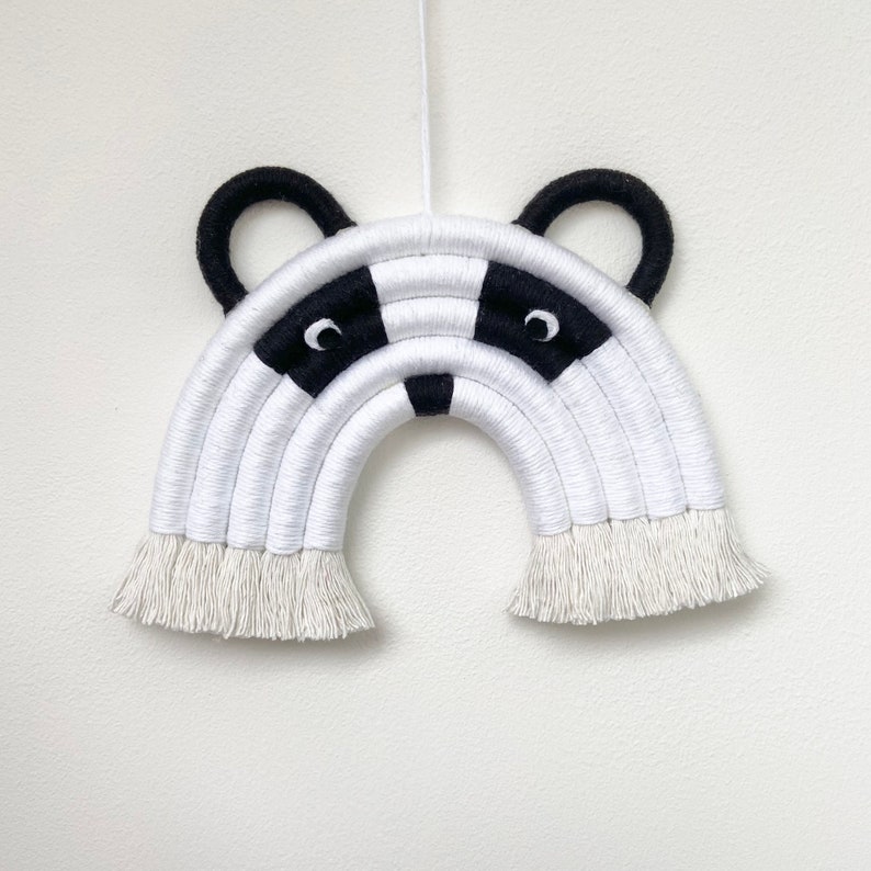 Puede incluir: Colgante de pared de arco&iacute;ris de macram&eacute; blanco y negro con un dise&ntilde;o de cara de tej&oacute;n. El arco&iacute;ris tiene una franja en la parte inferior.
