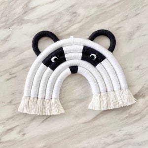 Puede incluir: Un tapiz de pared de arco&iacute;ris de macram&eacute; blanco y negro con una cara de mapache en el centro. El arco&iacute;ris est&aacute; hecho de hilo blanco con acentos de hilo negro. La cara de mapache tiene ojos negros y una nariz negra.