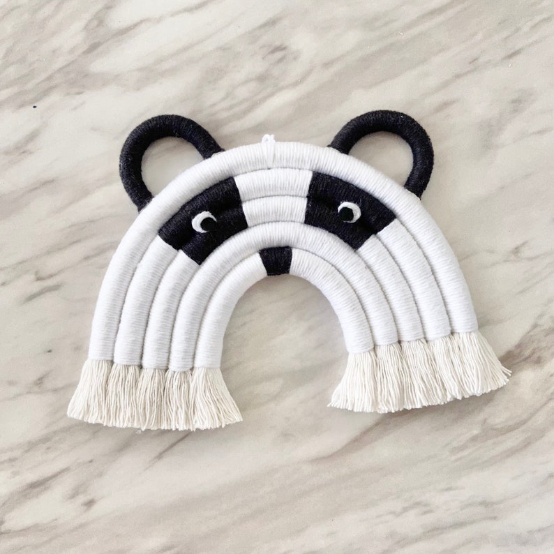 Puede incluir: Un tapiz de pared de arco&iacute;ris de macram&eacute; blanco y negro con una cara de mapache en el centro. El arco&iacute;ris est&aacute; hecho de hilo blanco con acentos de hilo negro. La cara de mapache tiene ojos negros y una nariz negra.