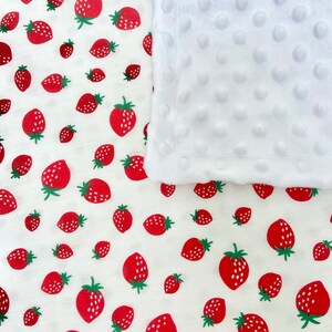 May include: A white baby blanket with a soft, textured surface and a red strawberry print. The blanket is made of two layers, a white fleece layer and a cotton layer with the strawberry print.