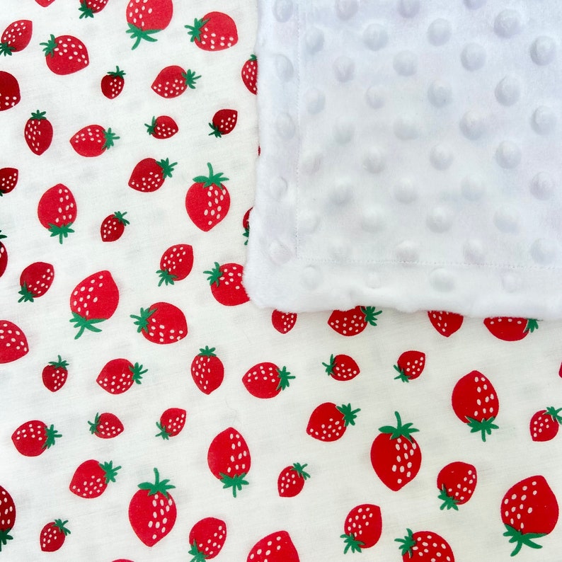 May include: A white baby blanket with a soft, textured surface and a red strawberry print. The blanket is made of two layers, a white fleece layer and a cotton layer with the strawberry print.