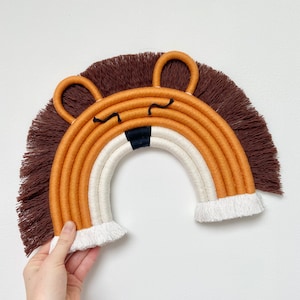 Puede incluir: Un arcoíris de pared marrón, naranja y blanco con una cara de león hecha de hilo marrón. El arcoíris está hecho de hilo grueso y tiene flecos en la parte inferior.