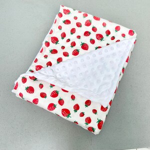 May include: A white and red baby blanket with a strawberry print on one side and a white, textured, soft fabric on the other side.