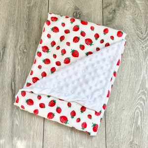 May include: A white and red baby blanket with a strawberry print. One side of the blanket is soft and fuzzy, while the other side is made of a smooth, cotton fabric.