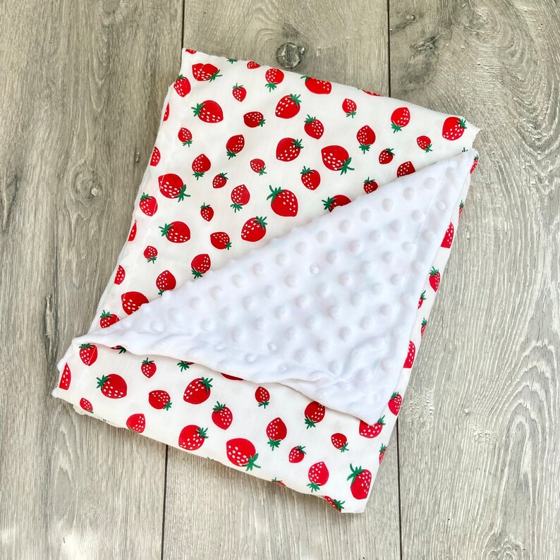 May include: A white and red baby blanket with a strawberry print. One side of the blanket is soft and fuzzy, while the other side is made of a smooth, cotton fabric.