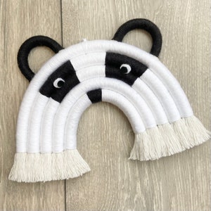 Puede incluir: Una decoraci&oacute;n de pared de arco&iacute;ris tejido en blanco y negro con un dise&ntilde;o de cara de panda. El arco&iacute;ris tiene flecos blancos y orejas negras.