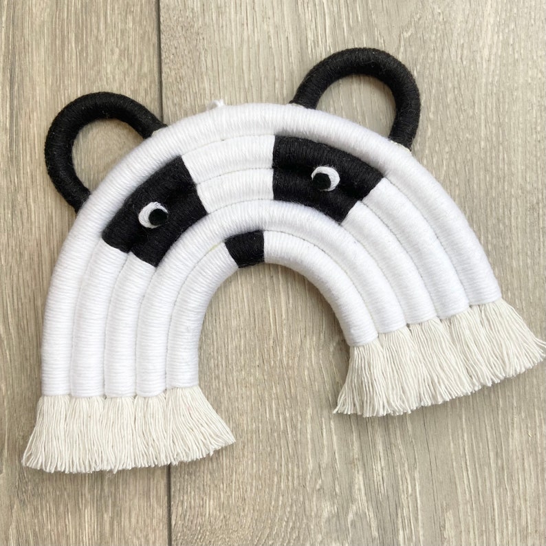Puede incluir: Una decoraci&oacute;n de pared de arco&iacute;ris tejido en blanco y negro con un dise&ntilde;o de cara de panda. El arco&iacute;ris tiene flecos blancos y orejas negras.