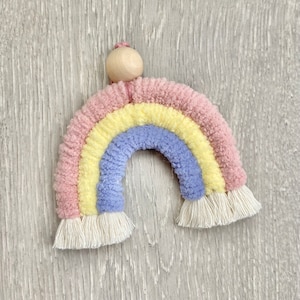 Może przedstawiać: Wiszący dekor ścienny w kształcie tęczy z różowej, żółtej i niebieskiej przędzy z drewnianym koralikiem na górze.