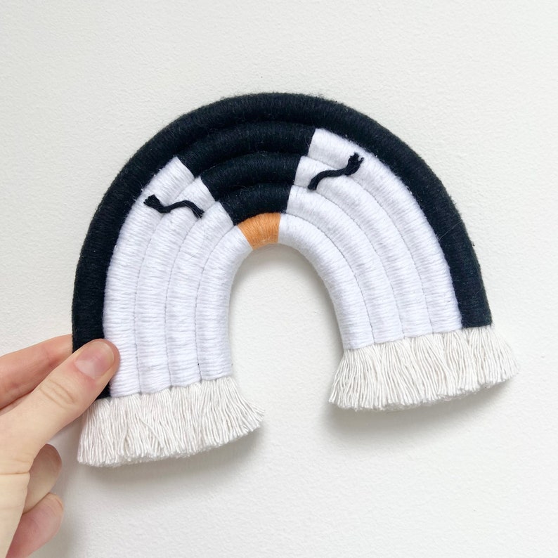 Op de afbeelding: Een zwart-witte macram&eacute; regenboog wandhanger met een franje rand. De regenboog heeft een klein oranje accent in het midden en twee zwarte ogen.