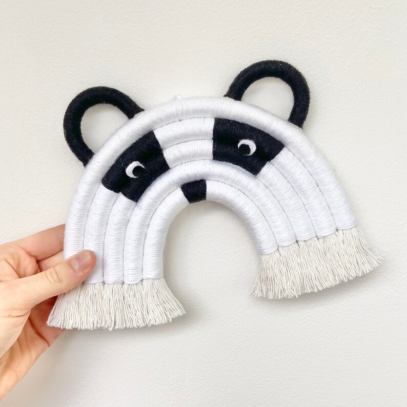 Puede incluir: Colgante de pared de arco&iacute;ris tejido en blanco y negro con un dise&ntilde;o de cara de mapache. El arco&iacute;ris tiene una franja en la parte inferior.