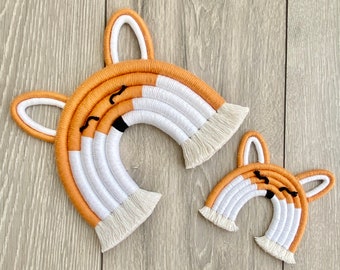 May include: Two macrame fox-shaped wall hangings. The larger one is approximately 10 inches wide, and the smaller one is about 5 inches wide. They feature orange, white, and black details, with a fringe at the bottom. The ears are arched and white.
