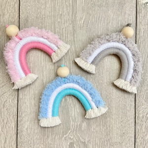 May include: Three handmade rainbow wall hangings. Each rainbow features a fluffy, textured top layer in pink, blue, and grey, with colourful arches and a wooden bead at the top. The rainbows are made with yarn and fringe.