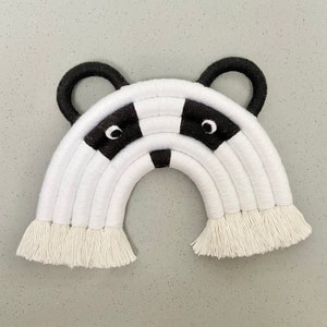 Puede incluir: Colgante de pared de arco&iacute;ris tejido en blanco y negro con una cara de mapache blanco y negro en el centro. El arco&iacute;ris tiene una franja en la parte inferior.